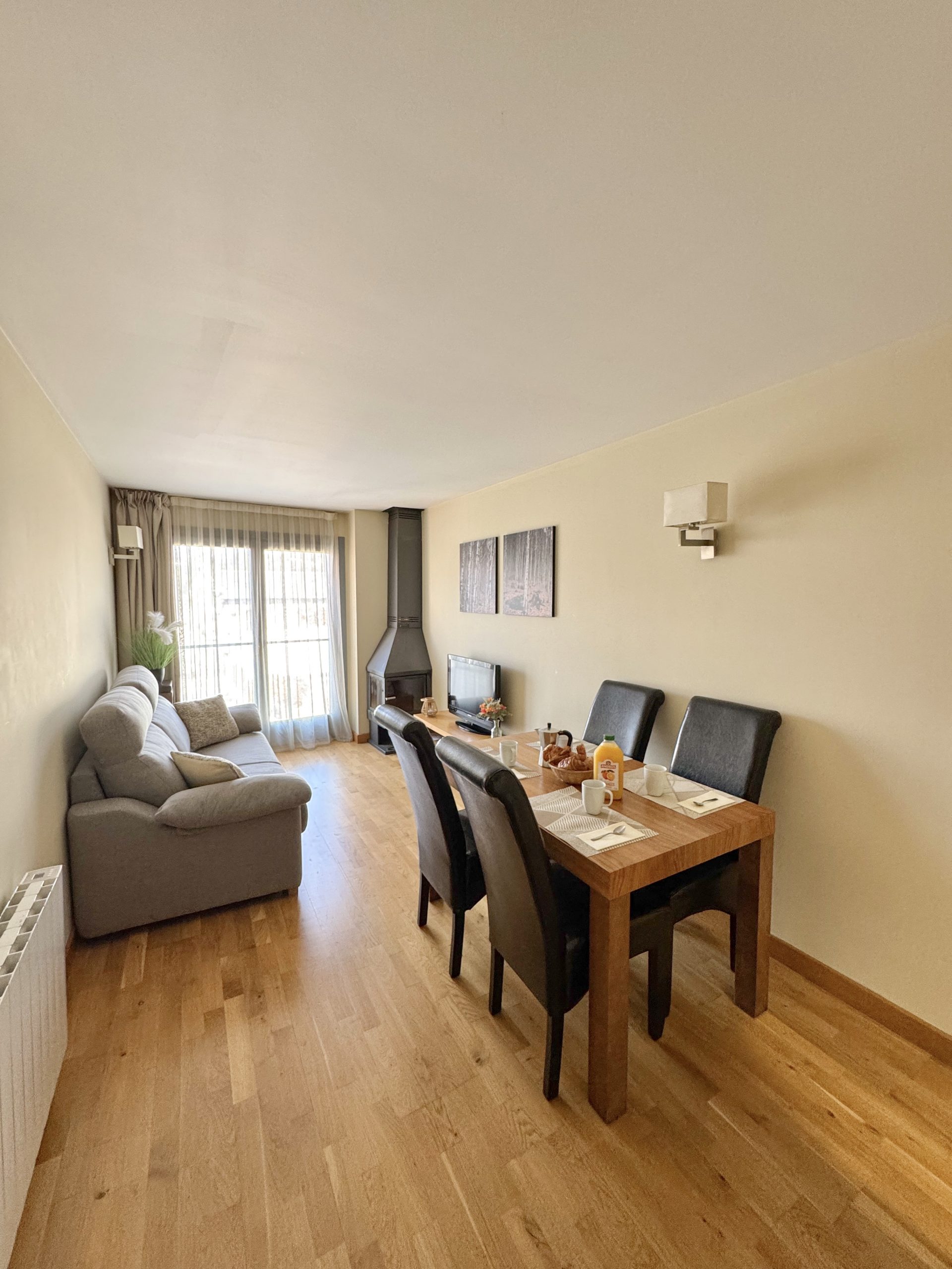 Salon lumineux avec canapé gris, coin repas en bois pour 4 personnes et poêle à bois, dans un appartement de l’Appart'hôtel Cerdanya.